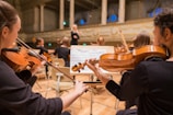 Musicians are seated in a grand concert hall, playing string instruments like violins. The conductor is visible in the background, leading the orchestra. The room is elegant, with high ceilings and ornate architectural details.