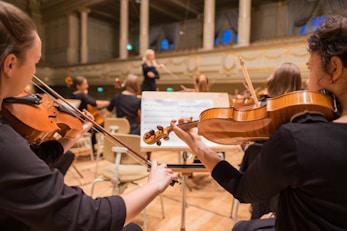 Musicians are seated in a grand concert hall, playing string instruments like violins. The conductor is visible in the background, leading the orchestra. The room is elegant, with high ceilings and ornate architectural details.