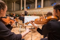 Musicians are seated in a grand concert hall, playing string instruments like violins. The conductor is visible in the background, leading the orchestra. The room is elegant, with high ceilings and ornate architectural details.
