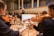 Musicians are seated in a grand concert hall, playing string instruments like violins. The conductor is visible in the background, leading the orchestra. The room is elegant, with high ceilings and ornate architectural details.