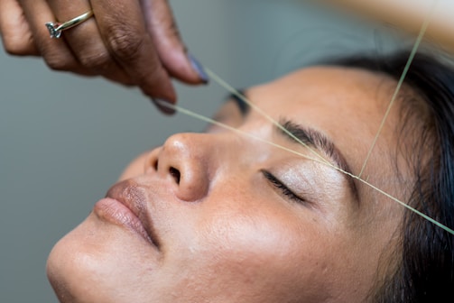 A close-up of a person receiving eyebrow threading. The person's eyes are closed, and a hand is expertly manipulating a thread across the eyebrows. The atmosphere is serene and focused.