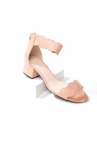 A stylish, peach-colored suede sandal with a block heel and ankle strap. The design features a unique scalloped edge on the strap, and the shoe has a modern, minimalist aesthetic.