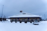 A Blockbuster Video store in a snowy environment, with the building covered in snow. The iconic blue and yellow signage stands out against the winter backdrop. There is a car parked in front of the building and an 'open' sign lit in red indicating the store is open for business.