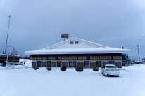 A Blockbuster Video store in a snowy environment, with the building covered in snow. The iconic blue and yellow signage stands out against the winter backdrop. There is a car parked in front of the building and an 'open' sign lit in red indicating the store is open for business.