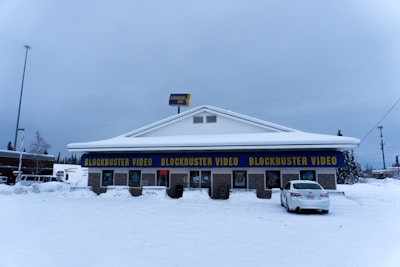 A Blockbuster Video store in a snowy environment, with the building covered in snow. The iconic blue and yellow signage stands out against the winter backdrop. There is a car parked in front of the building and an 'open' sign lit in red indicating the store is open for business.