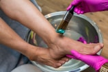 A person is receiving a foot spa treatment. The hands wearing pink gloves apply a teal-colored cream to the foot using a brush. The foot is positioned over a metallic basin filled with water.