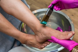 A person is receiving a foot spa treatment. The hands wearing pink gloves apply a teal-colored cream to the foot using a brush. The foot is positioned over a metallic basin filled with water.