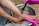 A person is receiving a foot spa treatment. The hands wearing pink gloves apply a teal-colored cream to the foot using a brush. The foot is positioned over a metallic basin filled with water.