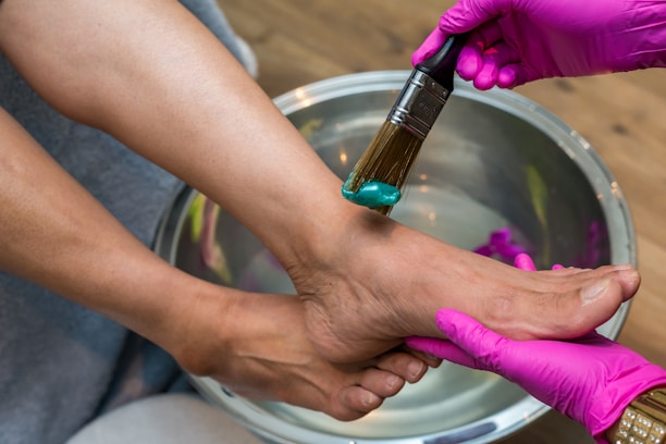 A person is receiving a foot spa treatment. The hands wearing pink gloves apply a teal-colored cream to the foot using a brush. The foot is positioned over a metallic basin filled with water.