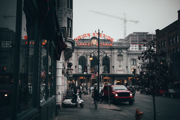 A foggy urban street scene featuring a historic train station with an illuminated sign reading 'Union Station Travel by Train'. The street is lined with buildings and construction cranes are visible in the background. A parked scooter and a red vehicle are seen in the foreground along with a few pedestrians.