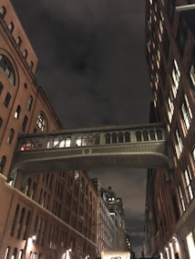 A dimly lit urban street scene featuring a bridge connecting two tall brick buildings. The buildings have multiple windows illuminated from within, creating a contrast with the dark, cloudy night sky. Streetlights cast a subtle glow on the sidewalk.