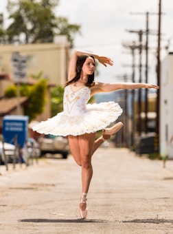 A ballet dancer performs an elegant pose on pointe, wearing a white tutu adorned with gold accents. The setting is an urban street with a blurred background, including buildings and cars.