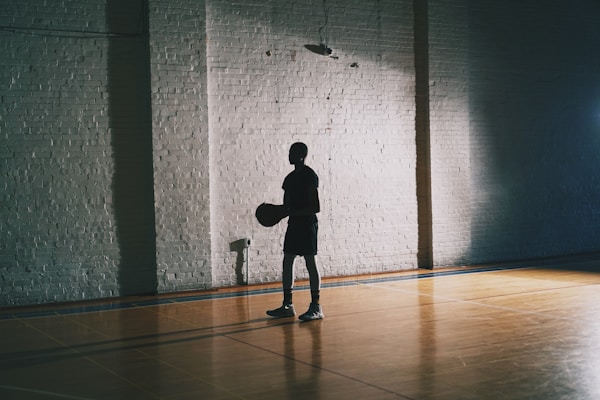 A silhouette of a person holding a basketball is cast against a brick wall inside an indoor basketball court. The wooden floor reflects light, and a shadow creates an atmospheric divide between light and darkness.
