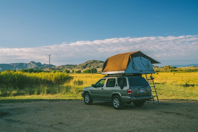 Silver SUV parked on a gravel lot beside a green field, a soft-shell rooftop tent unfolded on its roof with a tan rainfly and an aluminum ladder to the ground