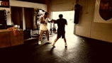 Two people are sparring in a dimly lit gym environment. The scene includes gym equipment such as a punching bag and exercise rig. One person is wearing boxing gloves and appears to be in an offensive stance, while the other is defensive. Natural light filters through an open garage door.