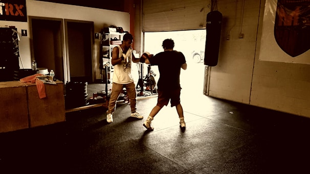 Two people are sparring in a dimly lit gym environment. The scene includes gym equipment such as a punching bag and exercise rig. One person is wearing boxing gloves and appears to be in an offensive stance, while the other is defensive. Natural light filters through an open garage door.