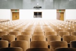 An empty lecture hall with rows of wooden seats facing a podium at the front. The room has a clock on the wall above the podium and exit doors on either side.