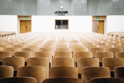 An empty lecture hall with rows of wooden seats facing a podium at the front. The room has a clock on the wall above the podium and exit doors on either side.