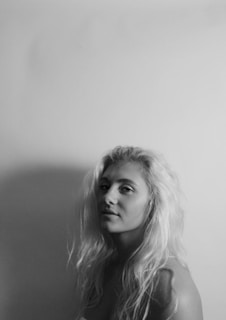A black and white portrait of a person with long hair gazing subtly towards the camera. The composition uses soft lighting against a plain background to create gentle shadows.