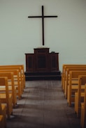 A simple church interior featuring a wooden cross mounted on a plain wall above a dark wooden pulpit. Yellow pews are aligned neatly on both sides of a wooden aisle, leading towards the altar.