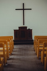 A simple church interior featuring a wooden cross mounted on a plain wall above a dark wooden pulpit. Yellow pews are aligned neatly on both sides of a wooden aisle, leading towards the altar.
