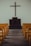 A simple church interior featuring a wooden cross mounted on a plain wall above a dark wooden pulpit. Yellow pews are aligned neatly on both sides of a wooden aisle, leading towards the altar.