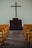A simple church interior featuring a wooden cross mounted on a plain wall above a dark wooden pulpit. Yellow pews are aligned neatly on both sides of a wooden aisle, leading towards the altar.
