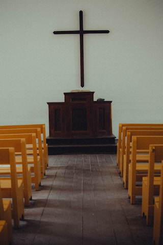 A simple church interior featuring a wooden cross mounted on a plain wall above a dark wooden pulpit. Yellow pews are aligned neatly on both sides of a wooden aisle, leading towards the altar.