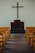 A simple church interior featuring a wooden cross mounted on a plain wall above a dark wooden pulpit. Yellow pews are aligned neatly on both sides of a wooden aisle, leading towards the altar.