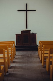 A simple church interior featuring a wooden cross mounted on a plain wall above a dark wooden pulpit. Yellow pews are aligned neatly on both sides of a wooden aisle, leading towards the altar.