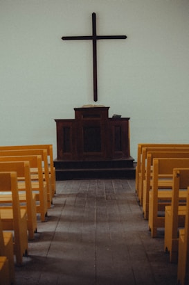A simple church interior featuring a wooden cross mounted on a plain wall above a dark wooden pulpit. Yellow pews are aligned neatly on both sides of a wooden aisle, leading towards the altar.