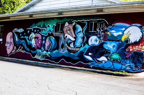 A vibrant mural featuring a series of interconnected images, including human faces, animals like an eagle and a whale, a turtle, and a landscape with mountains. The mural utilizes bold outlines and a mix of vivid colors, incorporating elements of nature alongside abstract motifs. It conveys cultural themes and possibly a narrative.