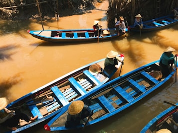 A group of individuals wearing traditional conical hats navigate several narrow wooden boats with vibrant blue accents along a calm, muddy river. The river is surrounded by dense, lush vegetation, and the lighting suggests a sunny day. The scene is peaceful, with reflections of the boats seen in the water.