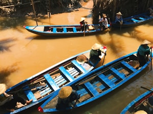 A group of individuals wearing traditional conical hats navigate several narrow wooden boats with vibrant blue accents along a calm, muddy river. The river is surrounded by dense, lush vegetation, and the lighting suggests a sunny day. The scene is peaceful, with reflections of the boats seen in the water.