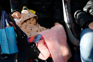 A young child is seated in a stroller, bundled up in a polka-dotted coat with a hood. The child's face peeks out, with only the eyes visible. A soft, pink blanket with a subtle pattern covers part of the stroller. Next to the stroller, a person wearing a black jacket is seated on what appears to be a bus. A blue tote bag with text and graphics is hanging from the stroller.