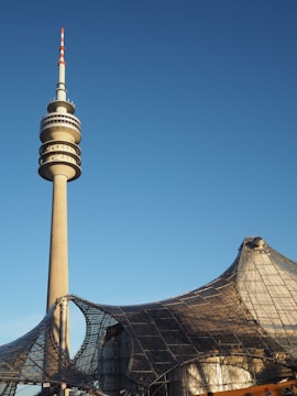 A tall, modern telecommunications tower stands prominently against a clear blue sky. To the right, a unique architectural structure features an intricate mesh design with curved and pointed elements, creating an impression of lightness and modernity.