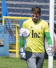 A soccer goalkeeper wearing a yellow jersey with a number 65, green gloves, and gray pants is seen training with a soccer ball. The ball is in mid-air close to the base of a goalpost. The background features a blue and yellow fence and a grassy area.