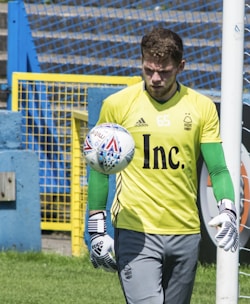 A soccer goalkeeper wearing a yellow jersey with a number 65, green gloves, and gray pants is seen training with a soccer ball. The ball is in mid-air close to the base of a goalpost. The background features a blue and yellow fence and a grassy area.