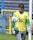A soccer goalkeeper wearing a yellow jersey with a number 65, green gloves, and gray pants is seen training with a soccer ball. The ball is in mid-air close to the base of a goalpost. The background features a blue and yellow fence and a grassy area.