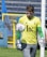 A soccer goalkeeper wearing a yellow jersey with a number 65, green gloves, and gray pants is seen training with a soccer ball. The ball is in mid-air close to the base of a goalpost. The background features a blue and yellow fence and a grassy area.