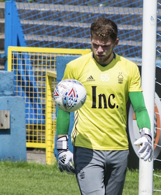 A soccer goalkeeper wearing a yellow jersey with a number 65, green gloves, and gray pants is seen training with a soccer ball. The ball is in mid-air close to the base of a goalpost. The background features a blue and yellow fence and a grassy area.