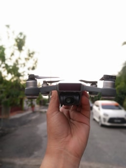 A person holds a small quadcopter drone with a camera in front, positioned prominently in the foreground. The background is a street with foliage on one side and a white car parked on the other, against a clear sky.