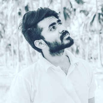A black and white portrait of a young man with a beard, looking slightly upward. He is wearing a light-colored polo shirt and has a thoughtful expression. The background features blurred palm trees, creating a serene outdoor setting.