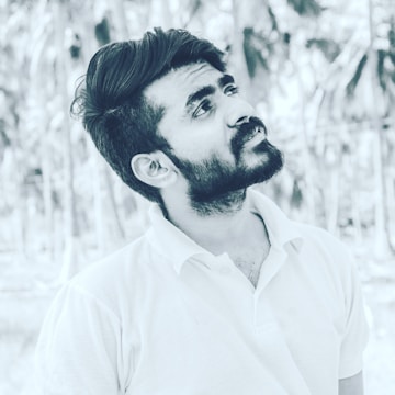 A black and white portrait of a young man with a beard, looking slightly upward. He is wearing a light-colored polo shirt and has a thoughtful expression. The background features blurred palm trees, creating a serene outdoor setting.