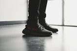 A pair of black sneakers with red lettering is worn by a person standing indoors on a polished floor near a large window. The shoes have a modern design with visible texture and detailed lacing.