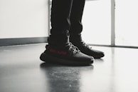 A pair of black sneakers with red lettering is worn by a person standing indoors on a polished floor near a large window. The shoes have a modern design with visible texture and detailed lacing.