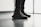 A pair of black sneakers with red lettering is worn by a person standing indoors on a polished floor near a large window. The shoes have a modern design with visible texture and detailed lacing.