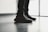 A pair of black sneakers with red lettering is worn by a person standing indoors on a polished floor near a large window. The shoes have a modern design with visible texture and detailed lacing.