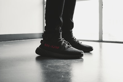 A pair of black sneakers with red lettering is worn by a person standing indoors on a polished floor near a large window. The shoes have a modern design with visible texture and detailed lacing.