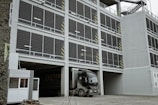 A multi-story parking garage with a large truck parked at the entrance. The facade consists of open metal grills and concrete, with safety markings on some pillars. Staircases are visible on the side of the structure, and there is a small security booth near the entrance.